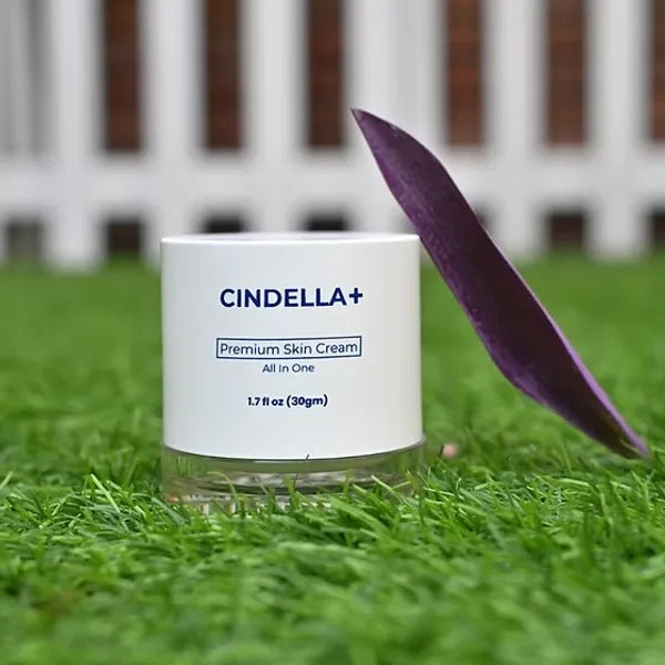 A white jar of CINDELLA+ Premium Skin Cream sits on green grass with a purple leaf leaning against it, and a blurred white fence is in the background.