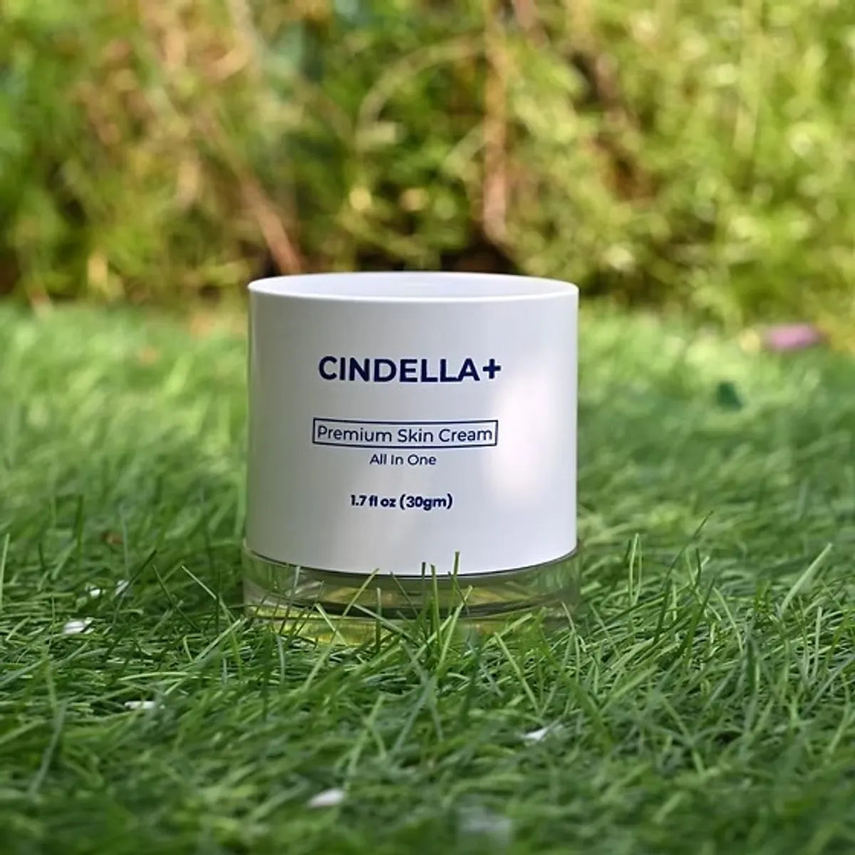 A white jar of CINDELLA+ Premium Skin Cream placed on green grass with a blurred natural background.
